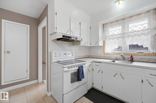 Kitchen featuring white cabinetry, a built-in range hood, a stainless steel sink, and light-colored tile flooring - 13304 63 Street Nw, Edmonton, AB 