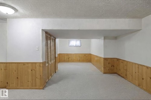 Spacious room featuring light-colored carpeting, wood-paneled wainscoting, white walls, and a textured ceiling.  This photo has been virtually decluttered. - 12035 83 Street Nw, Edmonton, AB 