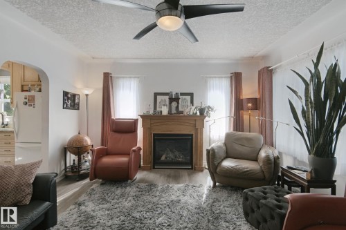 Bright living room anchored by a wood-burning fireplace with a decorative mantel, ceiling fan with integrated lighting, large windows with natural light, and neutral textured flooring throughout the open layout - 12035 83 Street Nw, Edmonton, AB 