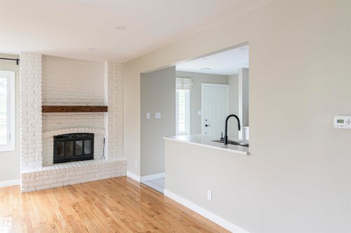 Living space featuring a whitewashed brick fireplace with a wood mantle, recessed lighting, wood-finish flooring, a pass-through window with a countertop, and a black gooseneck faucet - 5010 147 Street Nw, Edmonton, AB 