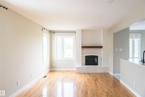 Spacious room featuring wood-finish flooring, a white-painted brick fireplace with a wood mantel, recessed lighting, and multiple windows - 5010 147 Street Nw, Edmonton, AB 
