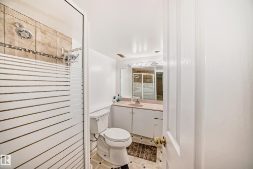 Shower with tiled walls and decorative accent band, frosted glass shower enclosure, vanity with built-in cabinetry, integrated sink, and wall-mounted mirror with overhead lighting, and patterned floor tiles - 4608 43 Avenue Nw, Edmonton, AB 