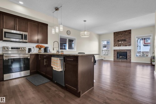 Open-concept living space featuring a kitchen island with pendant lighting, stainless steel appliances, dark wood-finish flooring, a stacked stone fireplace surround, and multiple windows - 3071 Carpenter Landing Sw, Edmonton, AB 