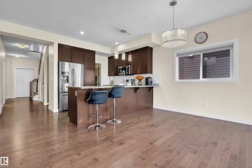 Open-concept kitchen and dining area featuring rich wood-finish flooring, dark wood cabinetry, a central island with a light-toned countertop, and stainless steel appliances - 3071 Carpenter Landing Sw, Edmonton, AB 