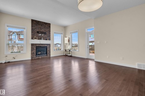 Spacious room featuring rich wood-finish flooring, a stone-clad fireplace with a white mantel, multiple windows, and a glass-paneled door opening to an exterior deck - 3071 Carpenter Landing Sw, Edmonton, AB 