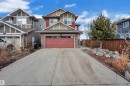Two-story exterior with red siding and light brown shake accents, featuring a two-car garage with a red door, a concrete driveway, and a rock garden with mature evergreen landscaping - 3071 Carpenter Landing Sw, Edmonton, AB 
