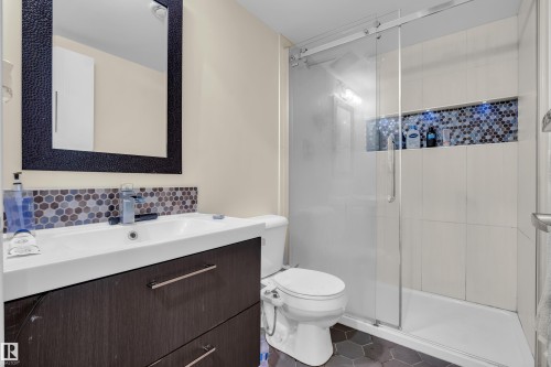 Contemporary bathroom featuring a dark wood-finish vanity with an integrated white sink, a mosaic tile backsplash, a frameless mirror with a textured black frame, a glass-enclosed shower with subway tile surround, and hexagonal floor tiles - 3071 Carpenter Landing Sw, Edmonton, AB 