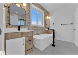 Freestanding soaking tub with a floor-mounted matte black faucet, dual vanities with wood-finish cabinetry and white countertops, and a full-height decorative tile accent wall - 