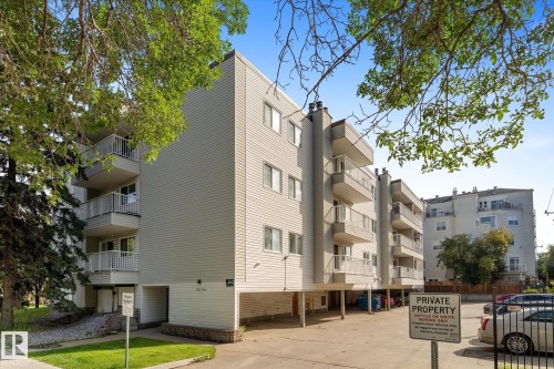 Multi-story building featuring light-colored siding, multiple balconies with white railings, and a ground-level parking area - #205 8125 110 St Nw, Edmonton, AB 