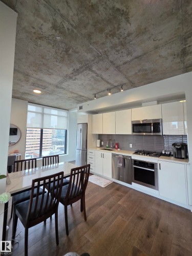 Industrial-style concrete ceiling, white flat-panel cabinetry, stainless steel appliances, wood-finish flooring, and a dining area with a window - #1006 1010 6 St Sw, Calgary, AB 