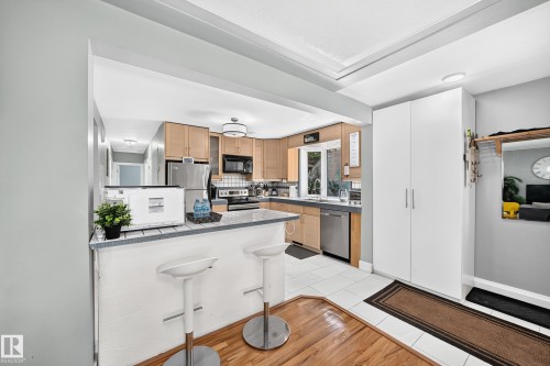 Kitchen with wood cabinetry, stainless steel appliances, a tiled breakfast bar, and white flooring - 10139 74 Street Nw, Edmonton, AB 