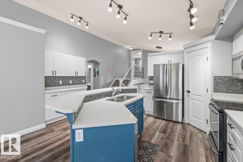 Kitchen featuring wood-finish flooring, white cabinetry, a contrasting blue island with a double basin sink, stainless steel appliances, and a mosaic tile backsplash - 15420 44 Street Nw, Edmonton, AB 