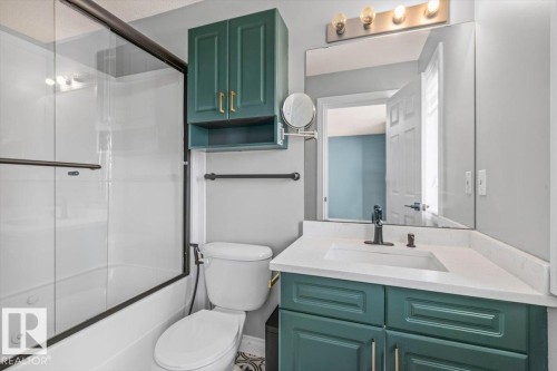 Shower-tub combination with dark-framed glass enclosure, teal cabinetry with gold-tone hardware, white countertop with integrated sink, and wall-mounted vanity lighting - 15420 44 Street Nw, Edmonton, AB 