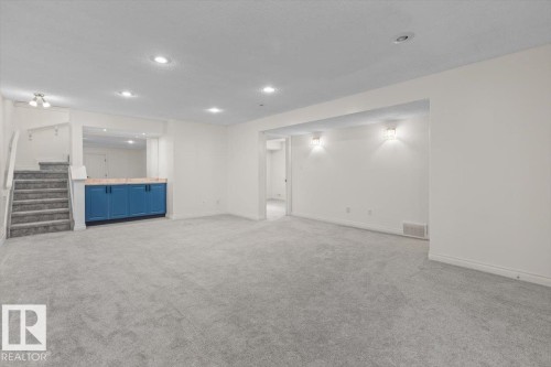 Spacious room featuring light gray carpeting, recessed ceiling lighting, a built-in cabinet with a wood-finish countertop, and a carpeted staircase - 15420 44 Street Nw, Edmonton, AB 