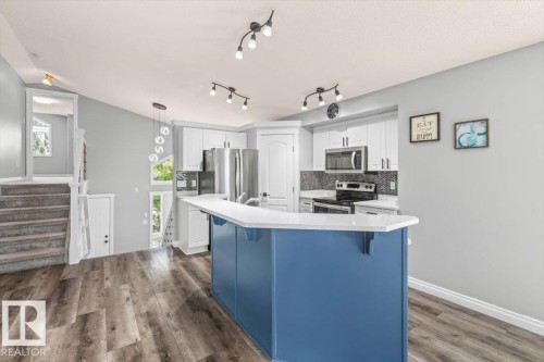 Modern kitchen with wood-finish flooring, a blue island with a white countertop, white cabinetry with stainless steel hardware, and a dark tile backsplash - 15420 44 Street Nw, Edmonton, AB 