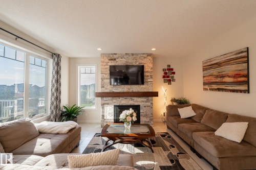Spacious living area featuring a stone-clad fireplace with a wood mantel, built-in recessed lighting, large windows, and light-toned flooring - 914 Crystallina Nera Way Nw, Edmonton, AB 