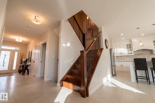 Wood-finish staircase with wrought iron balusters, white kitchen cabinetry, stainless steel appliances, mosaic tile backsplash, and light-toned tile flooring - 914 Crystallina Nera Way Nw, Edmonton, AB 