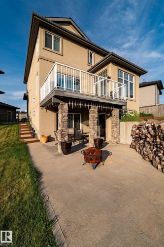 Two-story exterior with stucco siding, featuring a second-level deck with white railing, and a ground-level concrete patio with stone-clad support columns - 914 Crystallina Nera Way Nw, Edmonton, AB 