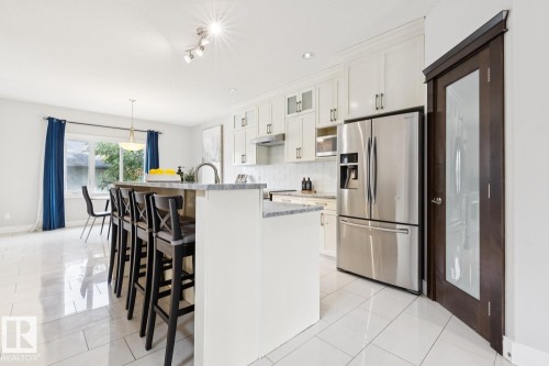 Expansive kitchen island with seating, stainless steel appliances, white cabinetry with display features, tiled flooring, and a glass-paneled interior door - 9919 79 Street Nw, Edmonton, AB 