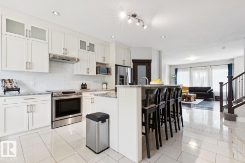 White cabinetry with brushed nickel hardware, stainless steel appliances, a center island with seating, a tile backsplash, and light-toned tile flooring - 9919 79 Street Nw, Edmonton, AB 