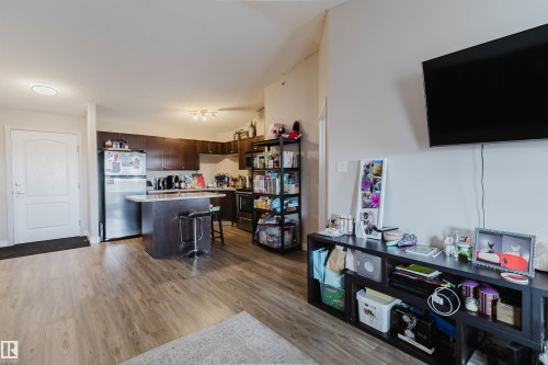 Open-concept living area with wood-finish flooring, a kitchen island with seating, stainless steel refrigerator, and dark wood cabinetry - #7428 7327 South Terwillegar Dr Nw, Edmonton, AB 