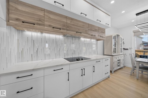 Kitchen featuring wood-finish upper cabinetry, white lower cabinetry, white backsplash with vertical accents, white countertops, and a built-in cooktop - 17128 2 Street Nw, Edmonton, AB 
