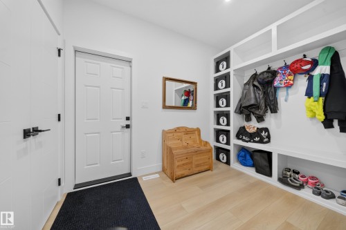 Entryway featuring light wood-finish flooring, white built-in storage with shelving and coat hooks, and a paneled entry door with black hardware - 17128 2 Street Nw, Edmonton, AB 