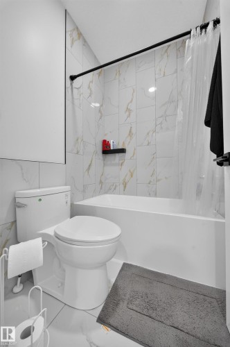 White bathtub with a black shower rod, white and gold-veined wall tiling, and a white toilet - 17128 2 Street Nw, Edmonton, AB 