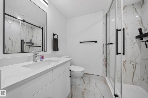 Full bathroom in basement with White vanity with an integrated sink and chrome faucet, large rectangular mirror with black frame, glass-enclosed shower with black fixtures and wall-mounted shelving, white and gold-veined tile flooring, and a white toilet - 17128 2 Street Nw, Edmonton, AB 
