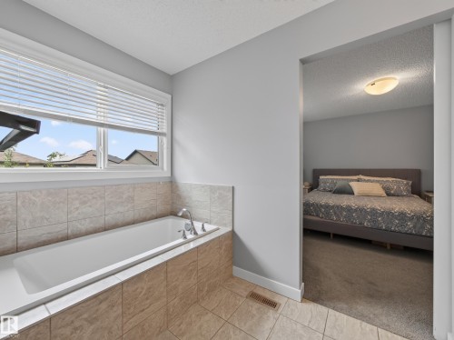 Bathroom featuring a built-in soaking tub with tile surround, a large window, and an open doorway to a carpeted room - 6504 48 Avenue, Beaumont, AB - Indoor Photo Showing Bathroom