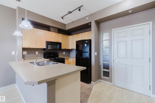 Kitchen featuring light wood cabinetry, black appliances, track lighting, and a kitchen island with integrated sink - #1-405 4245 139 Av Nw, Edmonton, AB 
