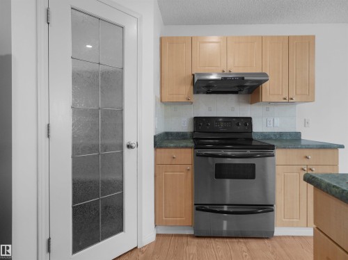 Stainless steel range with coordinating ventilation hood, wood-finish cabinetry, green countertops, white tiled backsplash, and a paneled glass door - 2410 32B Street Nw, Edmonton, AB 