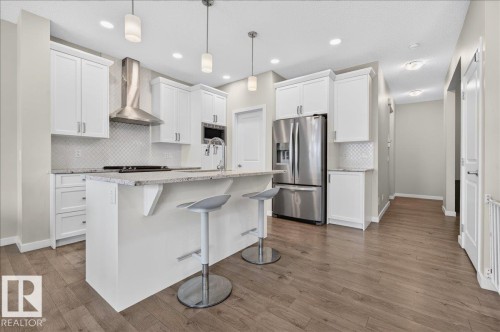 Kitchen featuring white cabinetry, stainless steel appliances, a center island with seating, a tile backsplash, and wood-finish flooring - 2709 Collins Crescent Sw Sw, Edmonton, AB - Indoor Photo Showing Kitchen With Stainless Steel Kitchen With Upgraded Kitchen