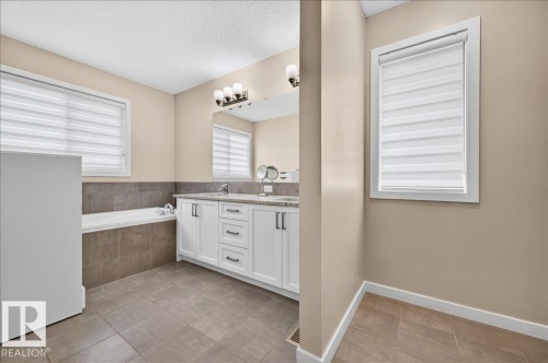 Bathroom featuring a built-in tub with tiled surround, dual vanity with undermount sinks and stone countertop, large mirror, and tiled flooring - 2709 Collins Crescent Sw Sw, Edmonton, AB - Indoor Photo Showing Bathroom