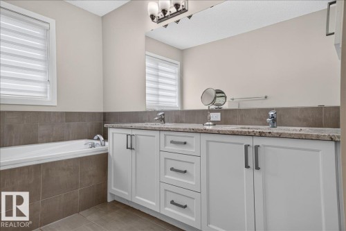 Bathroom featuring a dual-sink vanity with a light-toned countertop, white cabinetry with dark pulls, a built-in bathtub, and a window with horizontal blinds - 2709 Collins Crescent Sw Sw, Edmonton, AB - Indoor Photo Showing Bathroom