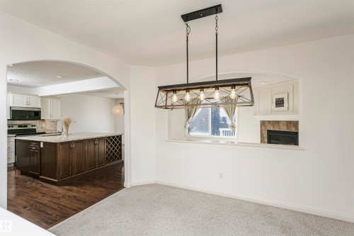 Open-concept living space featuring a carpeted area, a linear chandelier, a kitchen with a large island and wood-finish flooring, and a fireplace with a tiled surround - 2407 Hagen Way Nw, Edmonton, AB 