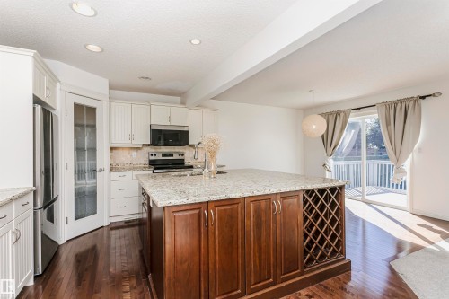 Kitchen featuring a large island with built-in wine storage, wood-finish flooring, white cabinetry, integrated stainless steel refrigerator, and a sliding glass door to an outdoor deck - 2407 Hagen Way Nw, Edmonton, AB 