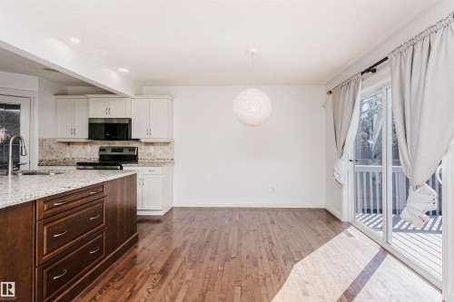 Wood-finish flooring throughout, kitchen island with stone countertop, white cabinetry with tile backsplash, stainless steel range and microwave, and sliding glass doors leading to a deck - 2407 Hagen Way Nw, Edmonton, AB 