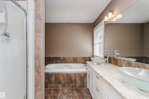 Dual vessel sinks set on a light-toned countertop, a built-in tub, a separate shower with a glass enclosure, and tiled flooring - 2407 Hagen Way Nw, Edmonton, AB 