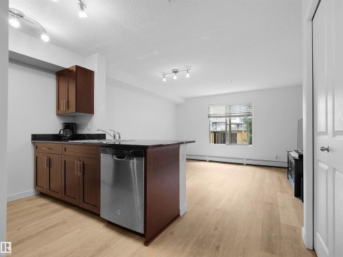 Kitchen island with integrated sink, stainless steel dishwasher, dark wood-finish cabinetry, and black countertops - #114 12035 22 Av Sw, Edmonton, AB 