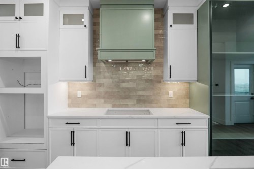 Contemporary kitchen featuring a muted green range hood, white shaker-style cabinetry with matte black hardware, stone-finish subway tile backsplash, and white countertops - 142 Edgewater Circle, Leduc, AB 