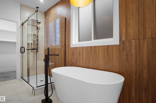 Freestanding soaking tub with a matte black floor-mounted faucet, wood-finish wall paneling, and a white framed window - 142 Edgewater Circle, Leduc, AB 