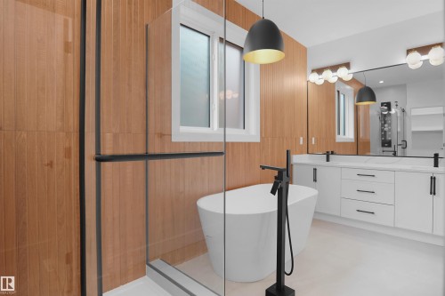 Freestanding soaking tub with a floor-mounted matte black faucet, wood-finish wall paneling, dual vanity with white cabinetry and integrated sinks, and a glass-enclosed shower - 142 Edgewater Circle, Leduc, AB 
