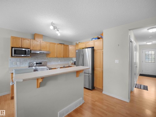 Kitchen featuring wood-finish cabinetry, stainless steel appliances, a light-toned backsplash, and an eat-at counter - 8111 7 Ave Avenue Sw, Edmonton, AB 