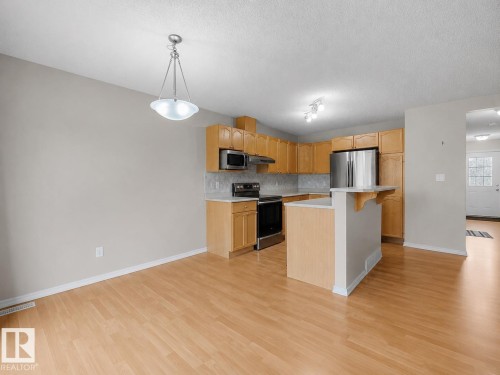 Open-concept kitchen featuring light wood cabinetry, stainless steel appliances, a breakfast bar, and wood-finish flooring - 8111 7 Ave Avenue Sw, Edmonton, AB 