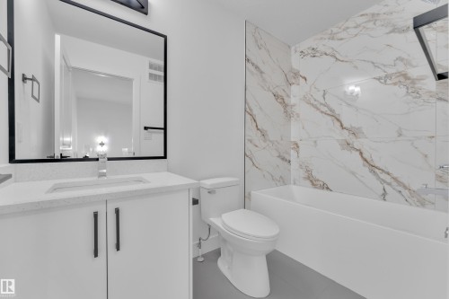 White vanity with a light-colored countertop, wall-mounted mirror with a black frame, and a built-in bathtub with a white and brown veined tile surround - 285 Munn Way, Leduc, AB 