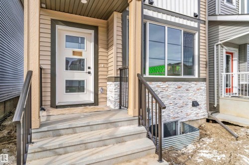 Entryway featuring a white door with rectangular glass inserts, concrete steps with dark railings, horizontal beige siding, and a stone veneer accent - 285 Munn Way, Leduc, AB 