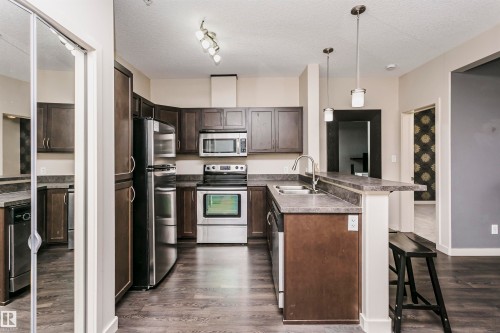 Kitchen featuring wood-finish flooring, stainless steel appliances, dark cabinetry, and a peninsula with a breakfast bar - #217 304 Ambleside Link Sw, Edmonton, AB 