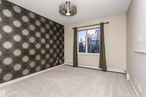Patterned accent wall, neutral wall paint, ceiling-mounted light fixture, window with drapes, and carpeted flooring - #217 304 Ambleside Link Sw, Edmonton, AB 