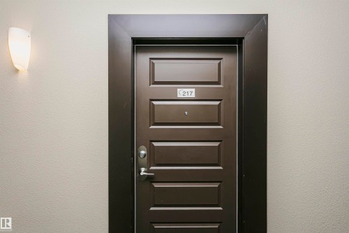 Paneled entry door with dark trim, featuring a wall-mounted sconce - #217 304 Ambleside Link Sw, Edmonton, AB 